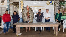 A imagem mostra um grupo de seis pessoas em pé atrás de uma mesa de madeira. No centro, há uma p...
