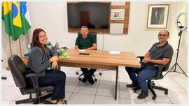 Três pessoas estão sentadas em uma sala de reuniões, em volta de uma mesa retangular de madeira....