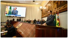Em um auditório oficial, um homem de terno escuro e gravata está em pé atrás de um púlpito, disc...