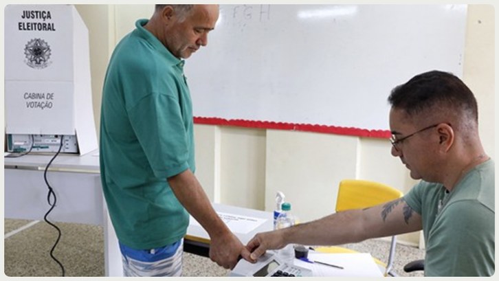 Atenção: prazo para regularizar título de eleitor termina segunda (19) Em uma sala de votação, dois homens interagem durante o processo eleitoral. Um homem de camisa v...