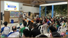 Uma mulher com roupa bege está a frente de vários alunos, ministrando uma palestra. Atrás dela, ...
