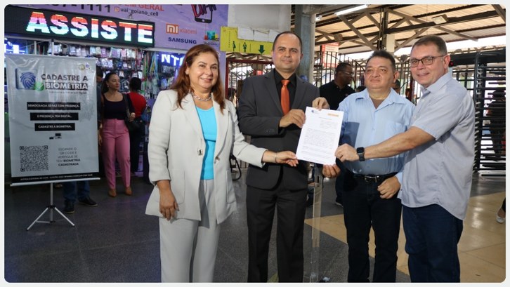 Quatro pessoas posam sorridentes segurando um documento em um ambiente movimentado. À esquerda, ...