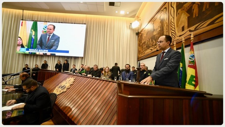 Em um auditório oficial, um homem de terno escuro e gravata está em pé atrás de um púlpito, disc...