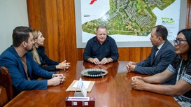 Cinco pessoas estão reunidas em volta de uma mesa de madeira em uma sala de reuniões. No centro,...
