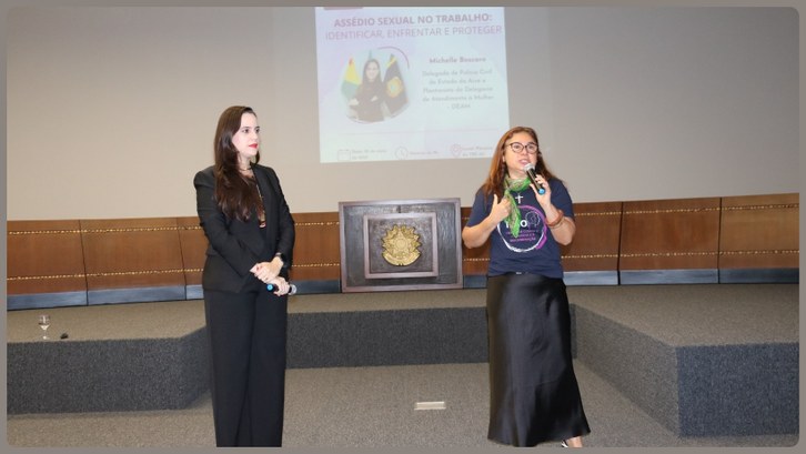 TRE-AC abre Semana de Combate ao Assédio com palestra sobre enfrentamento à violência contra a mulher Duas mulheres estão em pé sobre um palco durante uma palestra. À esquerda, uma mulher de terno p...