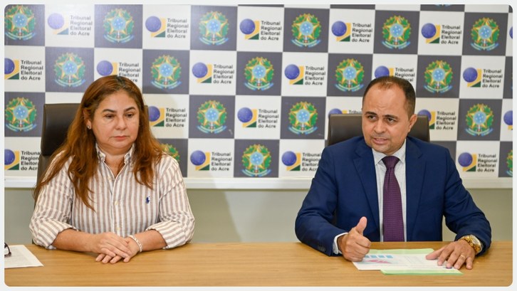 TRE-AC apresenta estratégias da Campanha da Biometria em coletiva de imprensa Duas pessoas estão sentadas lado a lado em uma mesa de reunião. À esquerda, uma mulher de cabelo...