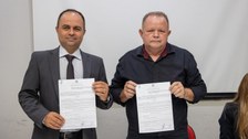 Dois homens estão lado a lado, de pé, segurando documentos assinados. O homem à esquerda usa ter...