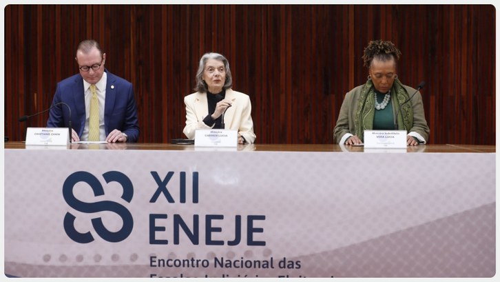 TRE-AC marca presença no XII Encontro Nacional das Escolas Judiciárias Eleitorais Três autoridades estão sentadas à mesa em um auditório com painel de madeira ao fundo. Ao centro...