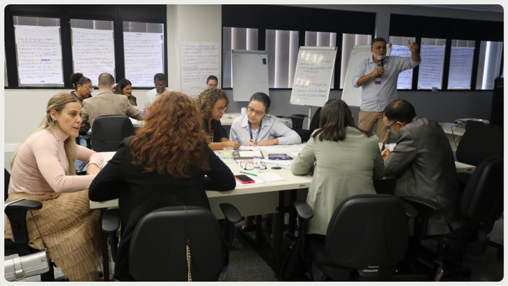 TRE-AC sedia curso para formação de laboratoristas de inovação no setor público Sala com cerca de 15 pessoas distribuídas em três grupos sentados ao redor de mesas retangulares...