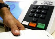 polegar no leitor biométrico com urna eletrônica ao fundo.